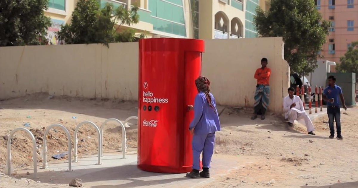 A red Coca-Cola “Hello Happiness” phone booth standing on a dusty street in Dubai, a migrant worker opening its door