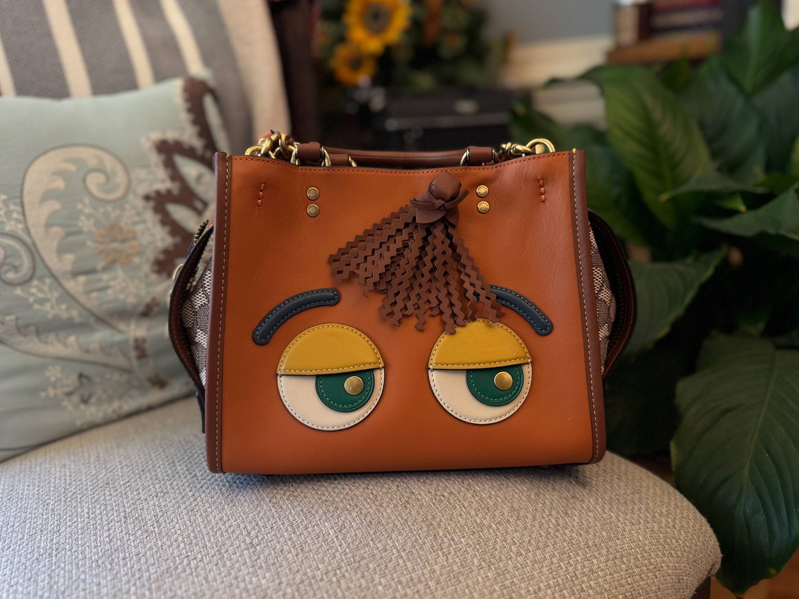 A tan leather Coach handbag with large Sesame Street style googly eyes and a leather fringe stitched onto the front, sitting on a woven chair