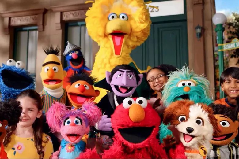 Big Bird, Elmo, Cookie Monster, Bert, Ernie, Abby and The Count gathered with a group of children on the Sesame Street stoop