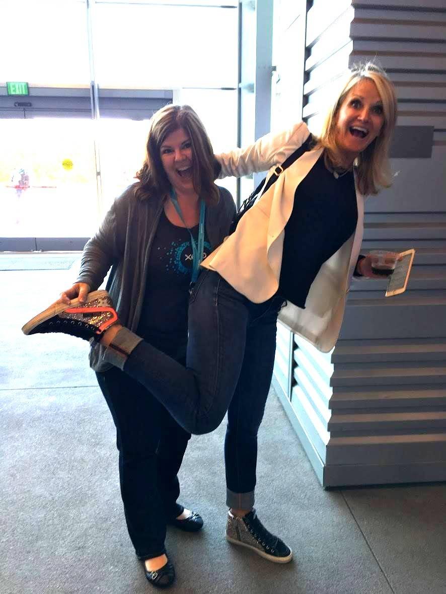 Jeannie Ruesch laughing with a colleague at a conference, holding up one of her studded sneakers