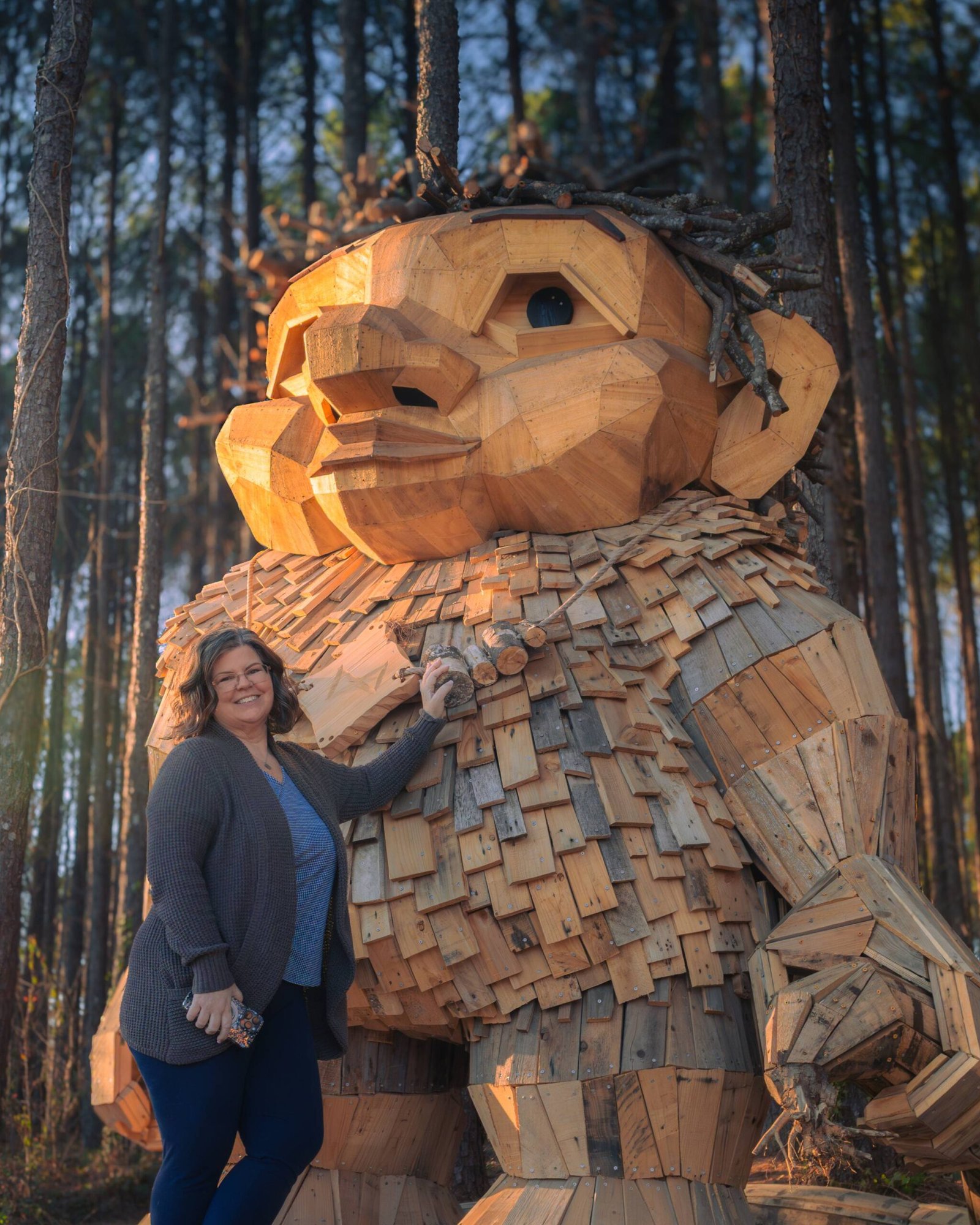 Jeannie Ruesch standing beside a giant troll sculpture built from reclaimed timber in a pine forest