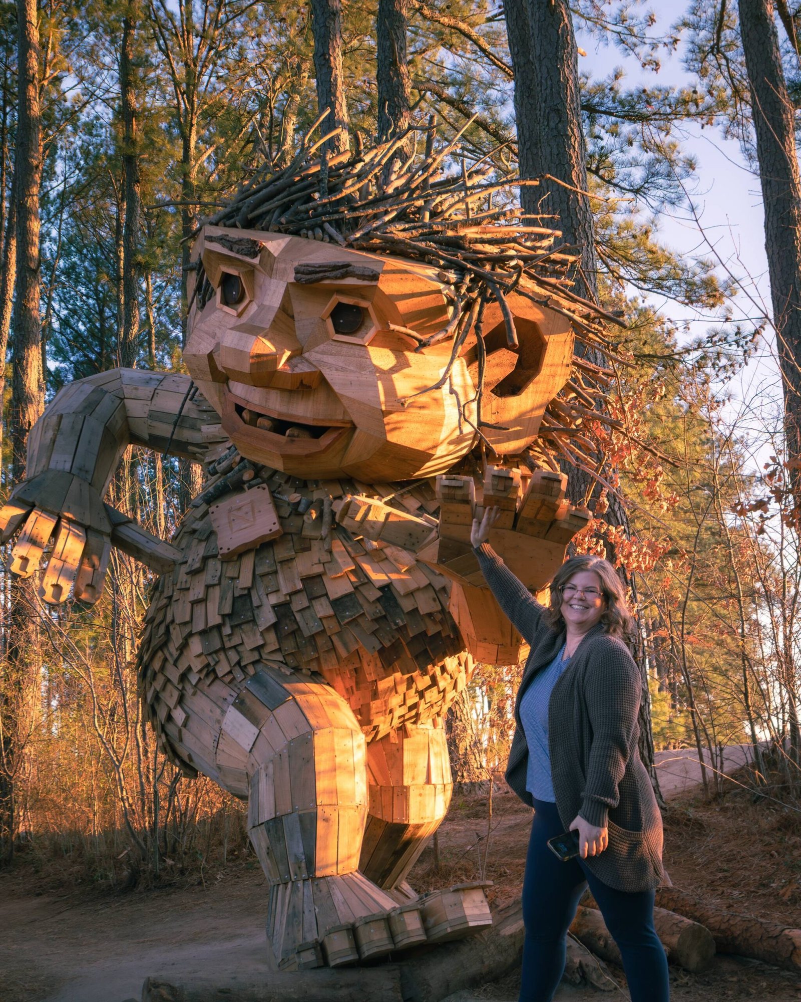Jeannie Ruesch waving beside a towering wooden troll sculpture with a crown of branches